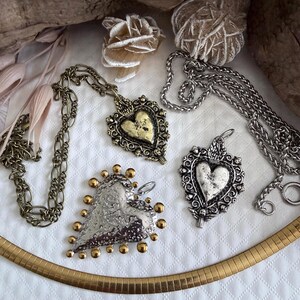 May include: A collection of vintage necklaces with heart-shaped pendants. The pendants have ornate designs in silver and gold tones, some with beaded accents. The necklaces are displayed on a white, textured surface, highlighting the intricate details of the jewellery.