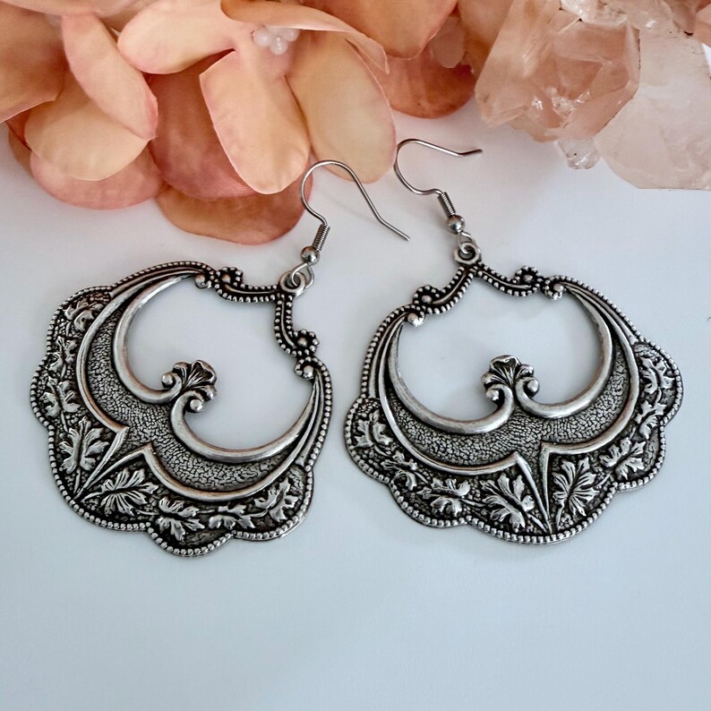 Pu&ograve; includere: Paio di orecchini argentati e ornati. Ogni orecchino presenta un elaborato motivo floreale e a volute all'interno di una grande forma traforata. Gli orecchini hanno una chiusura a gancio e sono esposti su uno sfondo morbido e neutro.