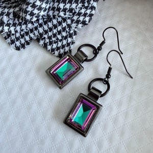 Peut inclure: Une paire de boucles d'oreilles rectangulaires avec des cristaux verts et violets irisés sertis dans des cadres en métal gris foncé. Les boucles d'oreilles ont une fermeture à crochet et un petit détail en anneau. Un tissu pied-de-poule noir et blanc est en arrière-plan.