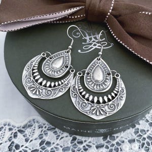 May include: Silver-coloured, ornate earrings with a teardrop and crescent design. The earrings feature intricate detailing and are suspended from a simple hook. They rest on a dark green box, accented by a brown ribbon.