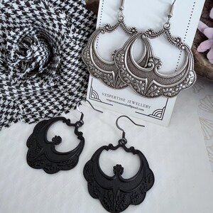 Pu&ograve; includere: Due paia di orecchini. Un paio &egrave; argentato con un design ornato e filigranato. L'altro paio &egrave; nero con un design simile, ma pi&ugrave; semplice. Gli orecchini sono esposti su una superficie bianca con un tessuto a fantasia bianco e nero sullo sfondo. Il testo "VESPERTINE JEWELLERY" &egrave; visibile.