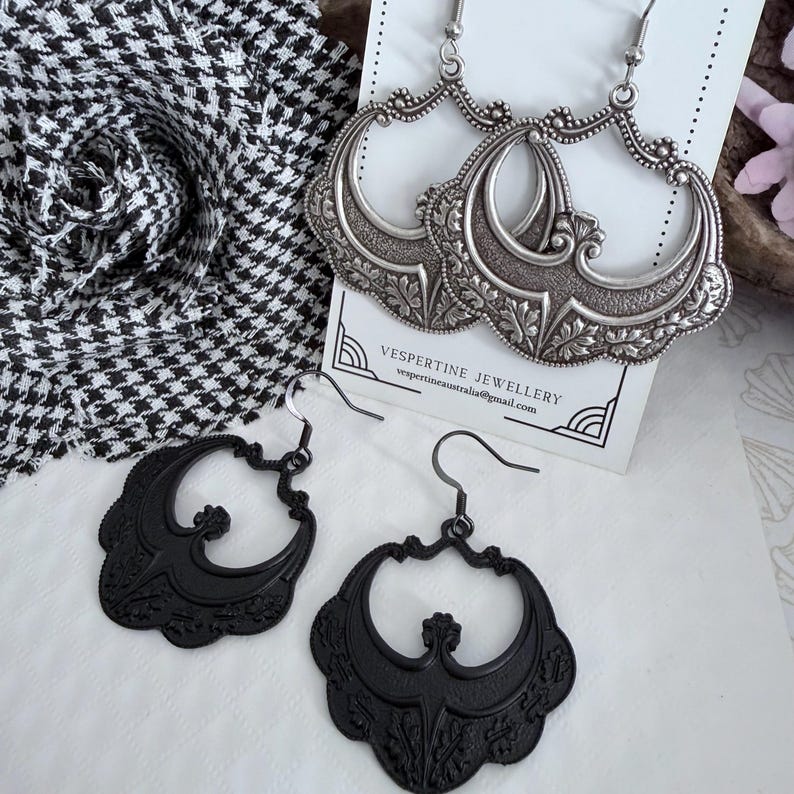 Pu&ograve; includere: Due paia di orecchini. Un paio &egrave; argentato con un design ornato e filigranato. L'altro paio &egrave; nero con un design simile, ma pi&ugrave; semplice. Gli orecchini sono esposti su una superficie bianca con un tessuto a fantasia bianco e nero sullo sfondo. Il testo "VESPERTINE JEWELLERY" &egrave; visibile.