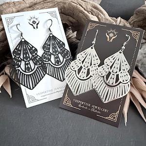 Peut inclure: Deux paires de boucles d'oreilles de style Art Déco. Une paire est noire, l'autre argentée. Les deux paires ont un design en forme d'éventail avec des motifs découpés complexes et sont présentées sur des cartes avec le texte "Vespertine Jewellery".