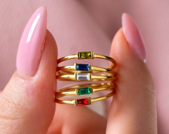 May include: Five gold rings with different colored gemstones. The rings are stacked on top of each other and are being held by a hand.