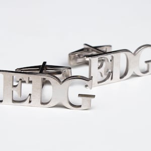 May include: Silver cufflinks with the letters "EDG" cut out. The cufflinks are on a white surface.