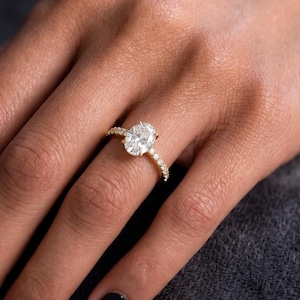 Puede incluir: Un anillo de oro con una gran piedra central de corte ovalado y una banda decorada con pequeñas piedras redondas. El anillo se lleva en un dedo, mostrando el engaste de metal pulido y el aspecto claro y facetado de la piedra preciosa.