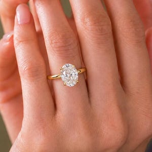 May include: A gold ring featuring a large, oval-cut, clear gemstone set in a four-prong setting. The band is a simple, polished gold metal. The oval stone is positioned centrally on the finger, showcasing a brilliant, faceted surface that reflects light.