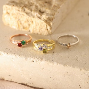 Pode incluir: Tr&ecirc;s an&eacute;is de ouro com pedras preciosas em forma de cora&ccedil;&atilde;o. Os an&eacute;is s&atilde;o adornados com pequenas pedras transparentes e apresentam pedras preciosas de diferentes cores: verde, roxo e laranja.