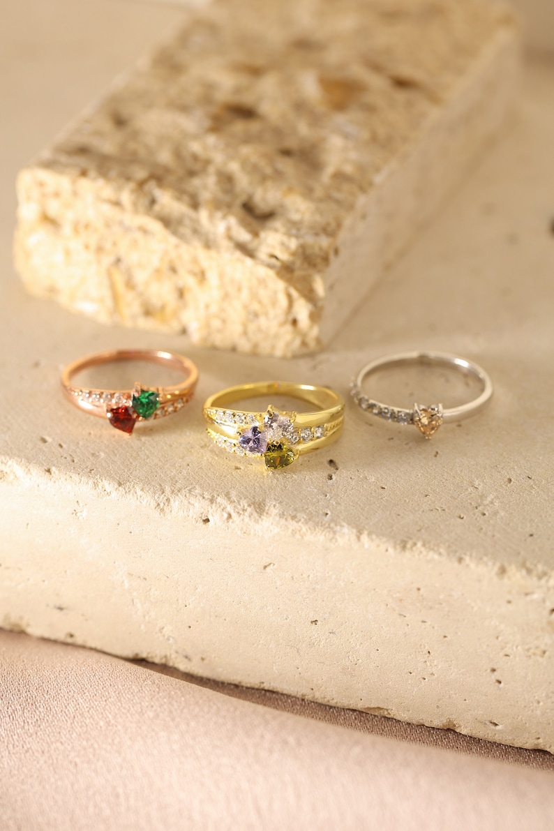 Pode incluir: Tr&ecirc;s an&eacute;is de ouro com pedras preciosas em forma de cora&ccedil;&atilde;o. Os an&eacute;is s&atilde;o adornados com pequenas pedras transparentes e apresentam pedras preciosas de diferentes cores: verde, roxo e laranja.