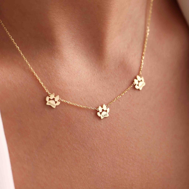 May include: A gold-colored necklace featuring three paw print charms. Each charm has a name engraved on it: "Martin", "Lexi", and another. The necklace is delicate and likely made of gold or a gold-colored metal.