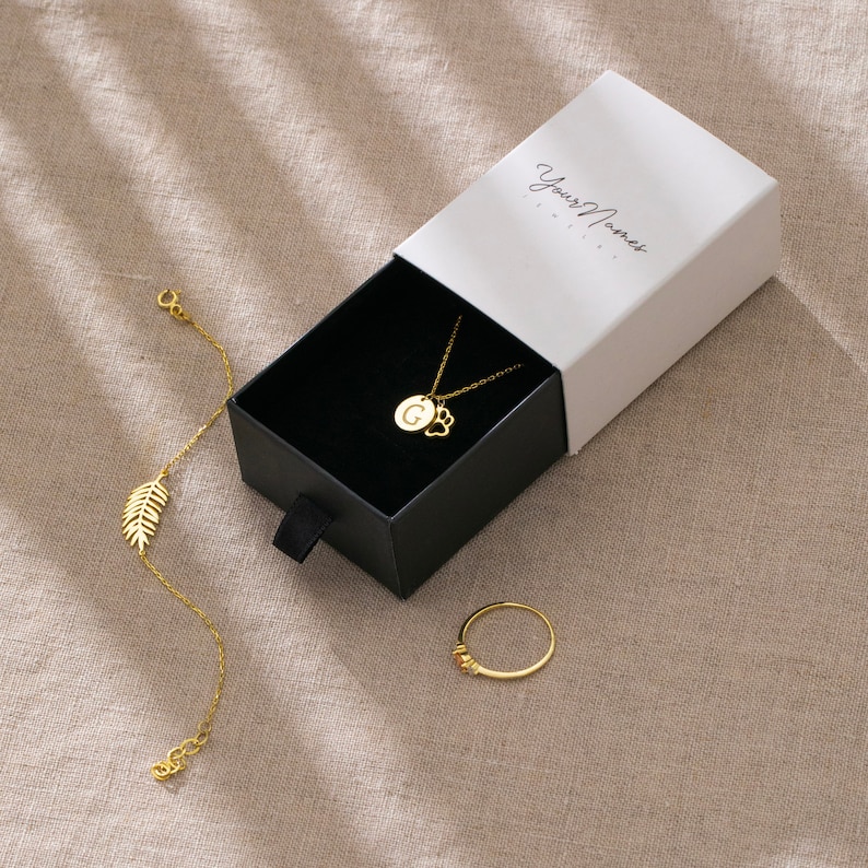 Pu&ograve; includere: Una collana d'oro con un ciondolo a forma di lettera G e un ciondolo a forma di impronta di zampa, un braccialetto d'oro con un ciondolo a forma di foglia e un anello d'oro con una piccola pietra. I gioielli sono in una scatola bianca con il testo "Your Moment Jewelry" sulla parte superiore.