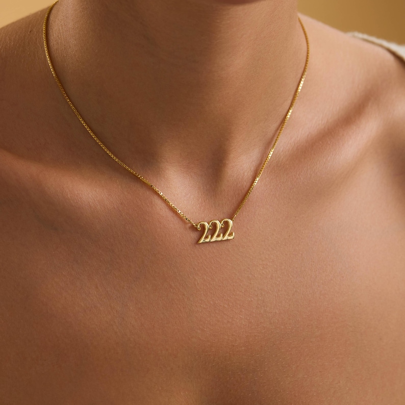 Pu&ograve; includere: Una collana color oro con un ciondolo con il numero "222". La collana, indossata intorno al collo, &egrave; composta da una catena sottile. Il ciondolo &egrave; in un carattere stilizzato.