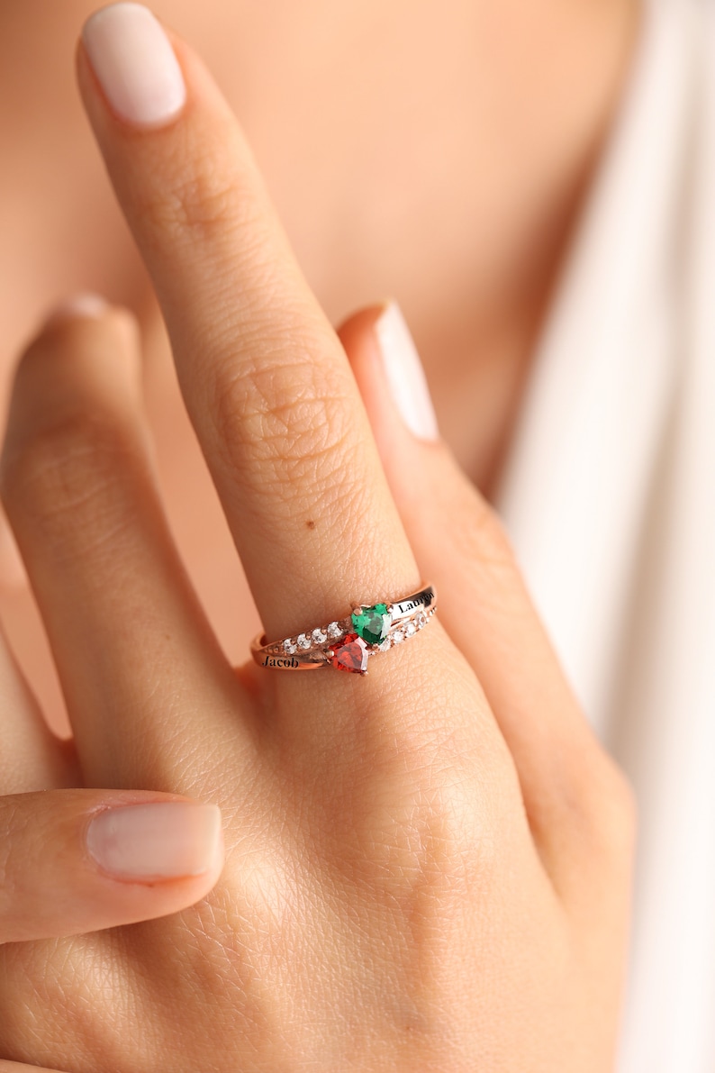 Pode incluir: Um anel de ouro rosa com duas pedras em forma de cora&ccedil;&atilde;o, uma vermelha e outra verde. O anel &eacute; gravado com os nomes "Jacob" e "Lacy".