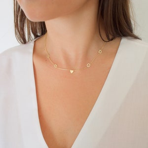 May include: A delicate gold necklace with a chain and several small, individual letters and a heart pendant. The letters are spaced along the chain. The necklace is worn against a white top.