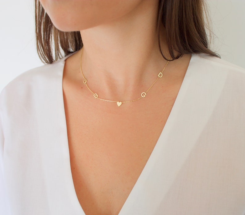 May include: A delicate gold necklace with a chain and several small, individual letters and a heart pendant. The letters are spaced along the chain. The necklace is worn against a white top.