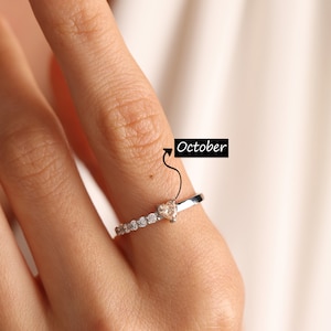 Pode incluir: Um anel de prata com uma pedra preciosa de top&aacute;zio em forma de cora&ccedil;&atilde;o, cercada por pedras preciosas menores e transparentes. O anel &eacute; usado em um dedo com esmalte de unha rosa claro. O texto "Outubro" est&aacute; escrito na imagem.