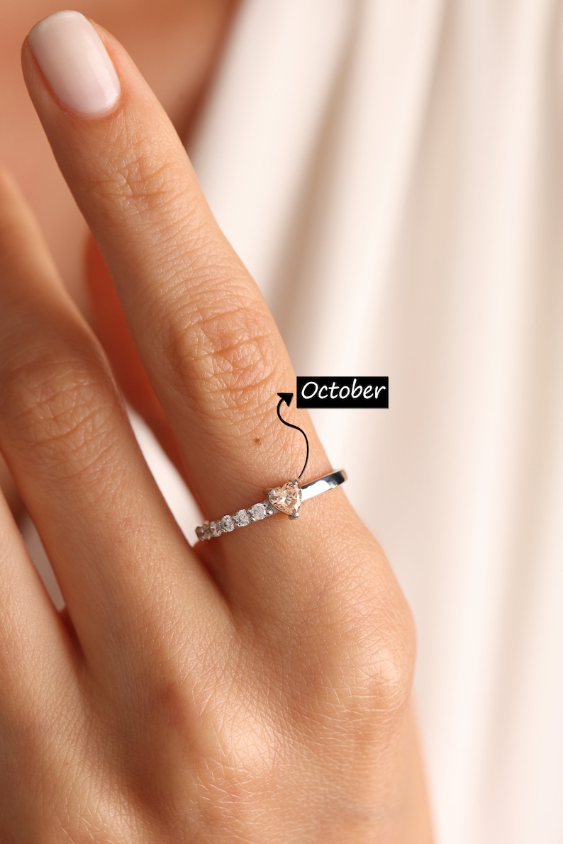Pode incluir: Um anel de prata com uma pedra preciosa de top&aacute;zio em forma de cora&ccedil;&atilde;o, cercada por pedras preciosas menores e transparentes. O anel &eacute; usado em um dedo com esmalte de unha rosa claro. O texto "Outubro" est&aacute; escrito na imagem.
