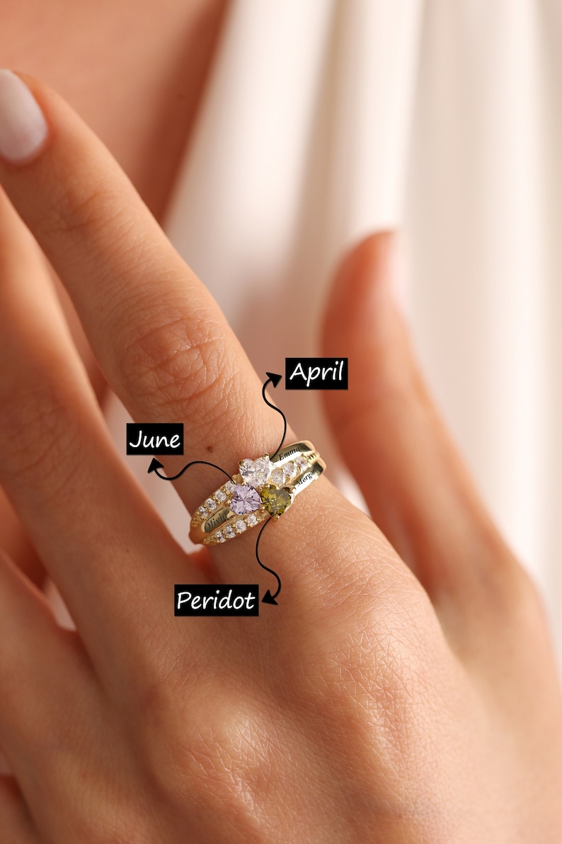 Pode incluir: Anel de ouro com tr&ecirc;s pedras em forma de cora&ccedil;&atilde;o. As pedras s&atilde;o de cor roxo claro, peridoto verde e transparente. O anel tem os nomes "Olivia", "Emma" e "Nora" gravados nele. O anel est&aacute; etiquetado como "Abril", "Junho" e "Peridoto".