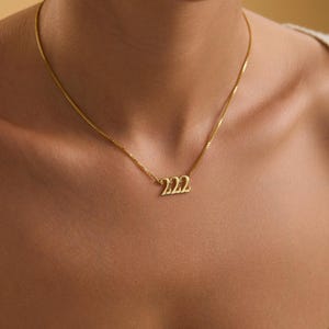 Pu&ograve; includere: Una collana color oro con una catena sottile. Il ciondolo presenta il numero "222" in un carattere stilizzato. La collana &egrave; indossata al collo di una persona, su uno sfondo neutro.