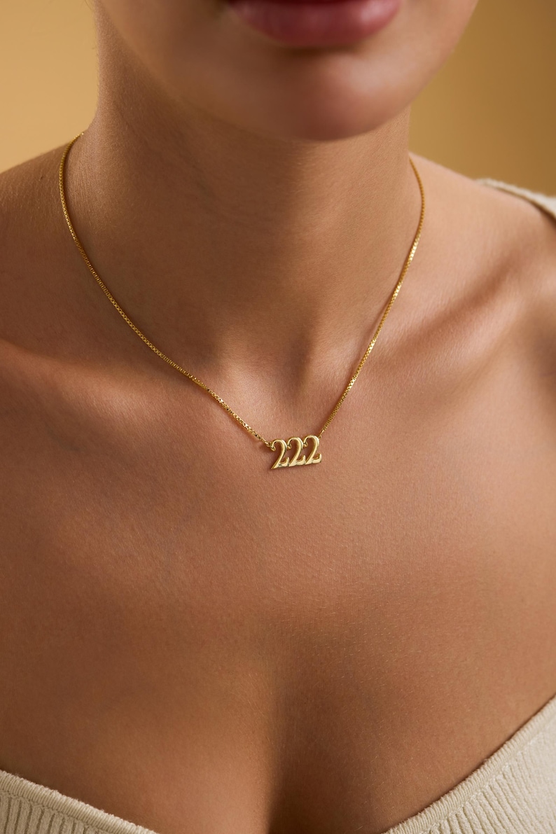 Pu&ograve; includere: Una collana color oro con una catena sottile. Il ciondolo presenta il numero "222" in un carattere stilizzato. La collana &egrave; indossata al collo di una persona, su uno sfondo neutro.