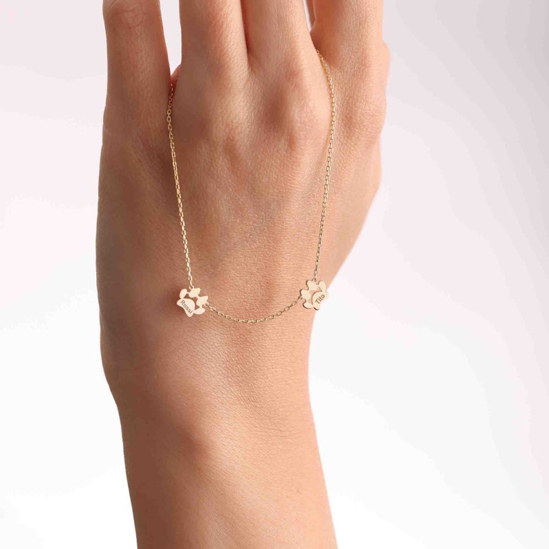 May include: A gold-colored necklace featuring three paw-shaped pendants. Each pendant has a name engraved on it. The necklace is displayed against a light background.