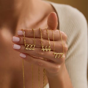 Pu&ograve; includere: Collane dorate con i numeri 222, 444 e 777 su catene sottili. Le collane sono tenute in mano, mostrando il design dei ciondoli e i dettagli delle catene. I gioielli sono esposti su uno sfondo neutro.