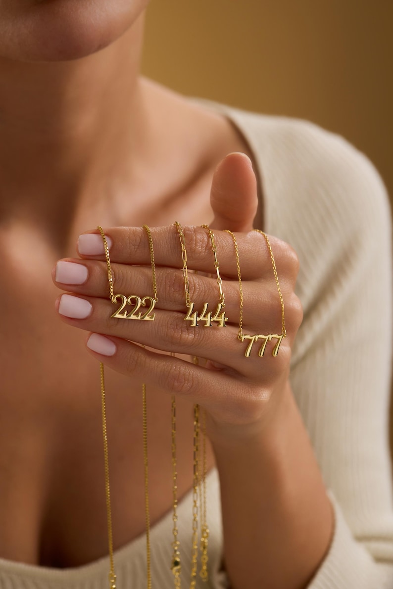 Pu&ograve; includere: Collane dorate con i numeri 222, 444 e 777 su catene sottili. Le collane sono tenute in mano, mostrando il design dei ciondoli e i dettagli delle catene. I gioielli sono esposti su uno sfondo neutro.