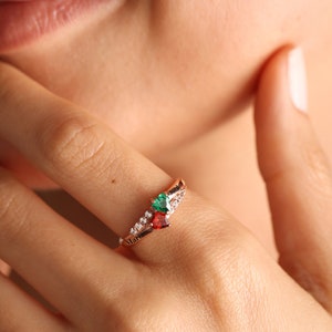 Pode incluir: Um anel de ouro rosa com uma gema vermelha e verde em forma de cora&ccedil;&atilde;o e uma banda de pequenas pedras claras. O anel tem a palavra "Maria" gravada na banda.