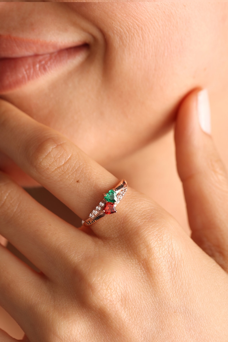 Pode incluir: Um anel de ouro rosa com uma gema vermelha e verde em forma de cora&ccedil;&atilde;o e uma banda de pequenas pedras claras. O anel tem a palavra "Maria" gravada na banda.