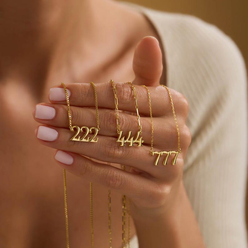Pu&ograve; includere: Collane dorate con i numeri 222, 444 e 777. I ciondoli sono sospesi a sottili catene. I gioielli sono tenuti in una mano con unghie dipinte, su uno sfondo sfocato.