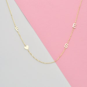 May include: A delicate gold-colored necklace with the letters "F", "E", and "S" spaced along the chain, along with a small heart charm. The necklace is displayed against a pink and gray background.