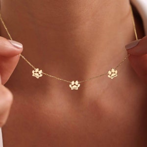 May include: A gold-colored necklace featuring three paw print charms. Each charm has a name engraved: "Merlin", "Lexi", and "Honey". The necklace is held by a person's hand, showcasing the delicate chain and pendant design.