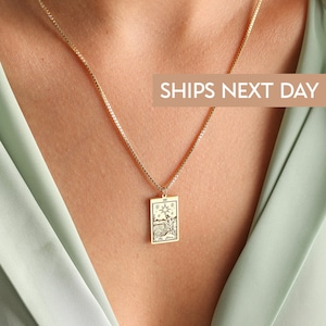 May include: A gold chain necklace with a rectangular pendant featuring a tarot card design. The card depicts a scene with a star, a figure, and water. The text "The Star" is visible on the card.