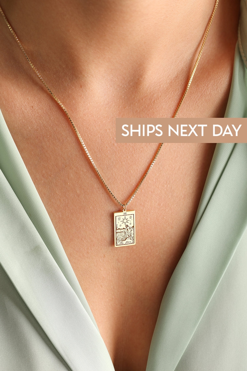 May include: A gold chain necklace with a rectangular pendant featuring a tarot card design. The card depicts a scene with a star, a figure, and water. The text "The Star" is visible on the card.