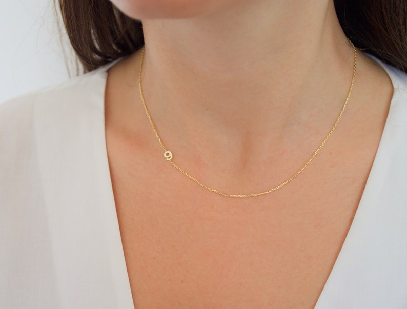 May include: A delicate gold-colored necklace featuring the number 9 pendant. The pendant is positioned to the side of the chain. The necklace is worn on a person's neck, against a white top.