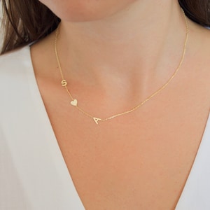 May include: A delicate gold necklace featuring the letters "S" and "A" with a small heart charm. The necklace is worn on a person with a white top. The jewelry is a personalized accessory.