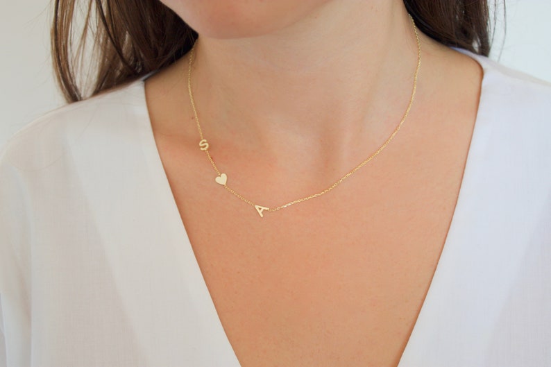 May include: A delicate gold necklace featuring the letters "S" and "A" with a small heart charm. The necklace is worn on a person with a white top. The jewelry is a personalized accessory.