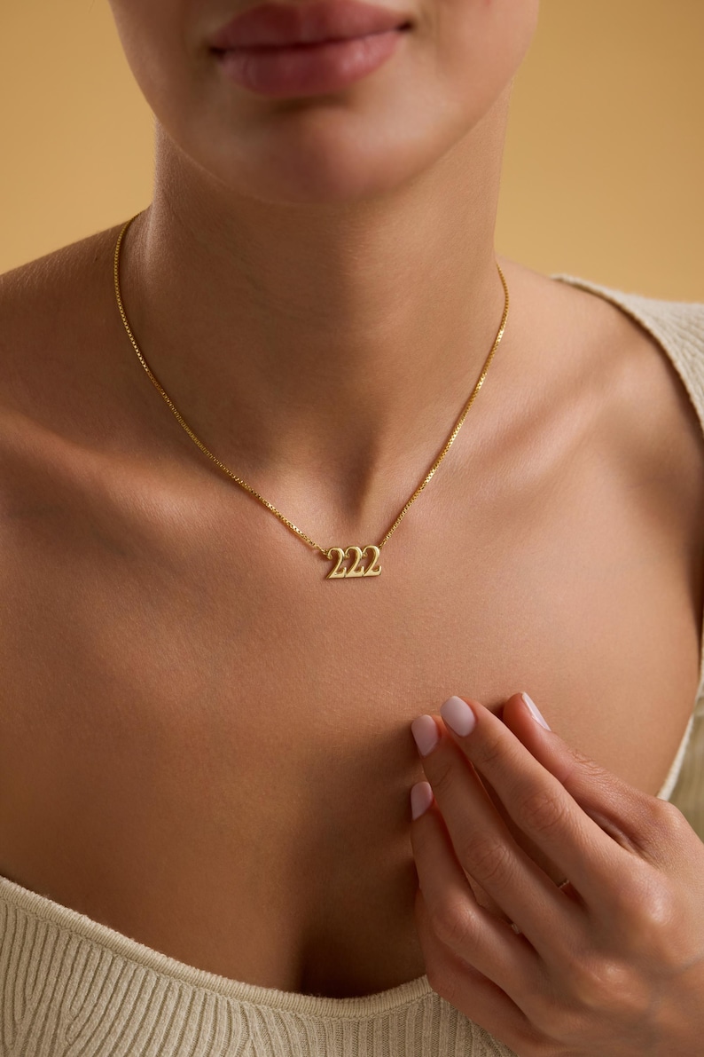 Pu&ograve; includere: Una collana color oro con un ciondolo con il numero "222". La collana &egrave; indossata intorno al collo. Lo sfondo &egrave; di colore beige.