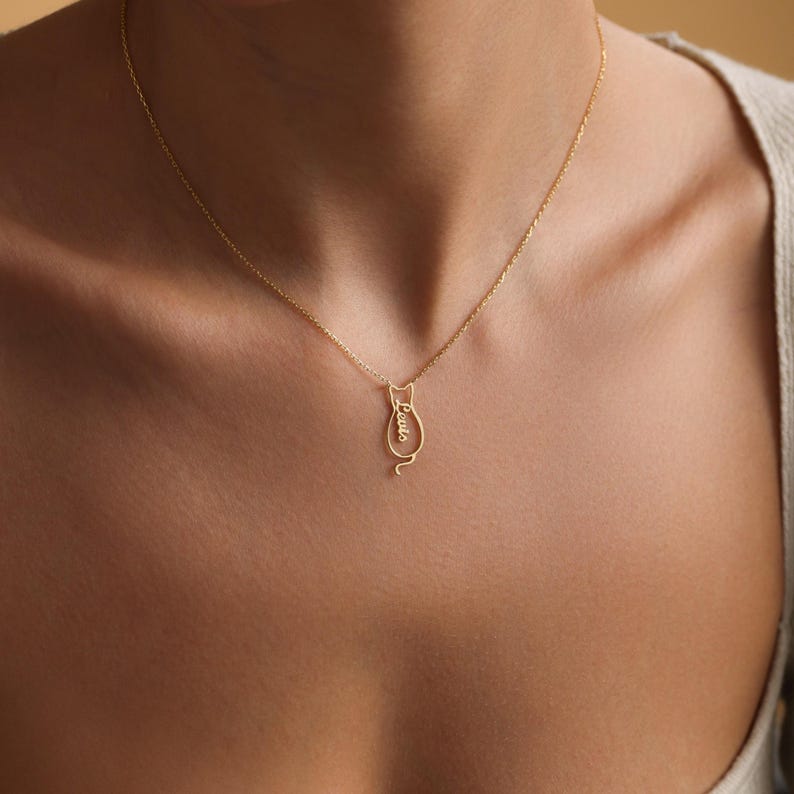 Puede incluir: Un collar de oro con un colgante con forma de gato. El colgante tiene la palabra "Lucas" escrita dentro del cuerpo del gato.