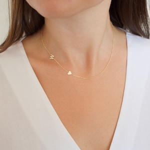 May include: A delicate gold necklace featuring a small letter "M" and a heart charm. The necklace is worn on a person, with the chain resting against a white top. The jewelry is simple and elegant.