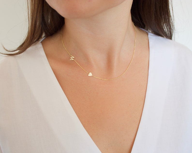 May include: A delicate gold necklace featuring a small letter "M" and a heart charm. The necklace is worn on a person, with the chain resting against a white top. The jewelry is simple and elegant.