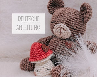 May include: A brown crocheted teddy bear with a white face and a red crocheted mushroom with a white stem and a brown heart button. The text "DEUTSCHE ANLEITUNG" is visible in the background.