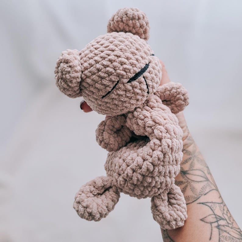 May include: A crocheted teddy bear in a soft, muted pink color. The bear has closed eyes with black stitched details, a small nose, and a rounded head. The plush toy has a textured, chunky knit appearance, with small arms and legs.