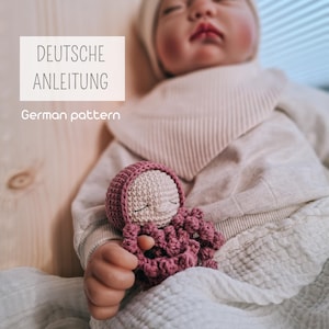 Könnte beinhalten: Handgefertigte Häkelpuppe in Rosa- und Cremetönen. Die Puppe hat ein schlafendes Gesicht und trägt eine passende Mütze. Der Text "DEUTSCHE ANLEITUNG" und "German pattern" ist im Bild enthalten.