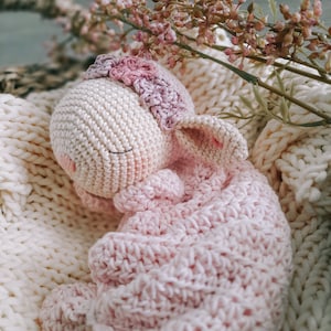 Op de afbeelding: Een gehaakt roze en wit knuffeldier in de vorm van een konijn, liggend in een witte gehaakte mand met roze bloemen.