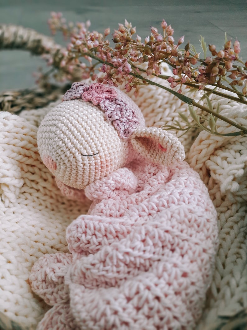 Op de afbeelding: Een gehaakt roze en wit knuffeldier in de vorm van een konijn, liggend in een witte gehaakte mand met roze bloemen.
