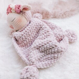Op de afbeelding: Een roze gehaakt knuffeldier met een bloemenkrans, gewikkeld in een roze gehaakt deken.