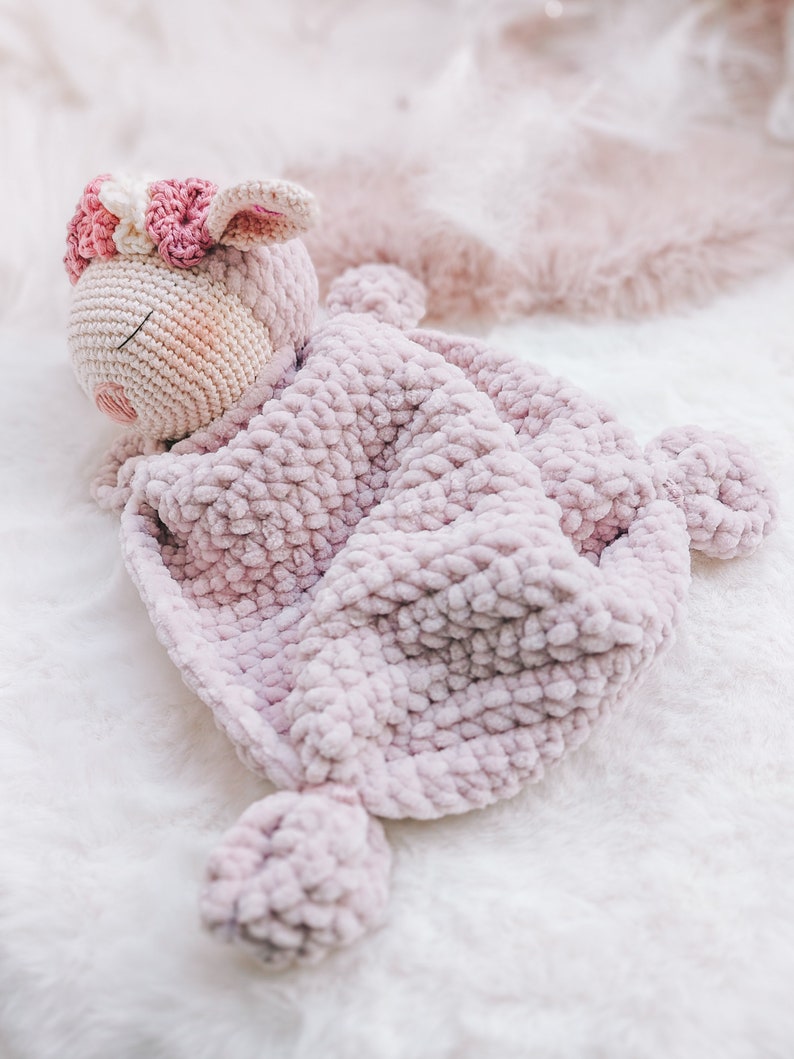 Op de afbeelding: Een roze gehaakt knuffeldier met een bloemenkrans, gewikkeld in een roze gehaakt deken.