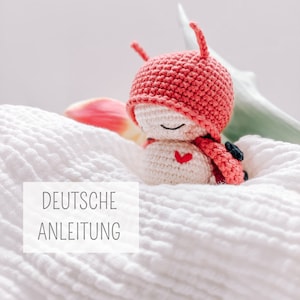 Puede incluir: Un juguete de mariquita de crochet con un sombrero rojo y un cuerpo blanco. El juguete tiene un pequeño corazón rojo en el pecho y está acostado sobre una manta blanca. El texto "DEUTSCHE ANLEITUNG" es visible en la imagen.