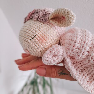 Op de afbeelding: Een gehaakt roze en wit lammetje met een roze deken. Het lammetje heeft een gesloten oog en een kleine mond.